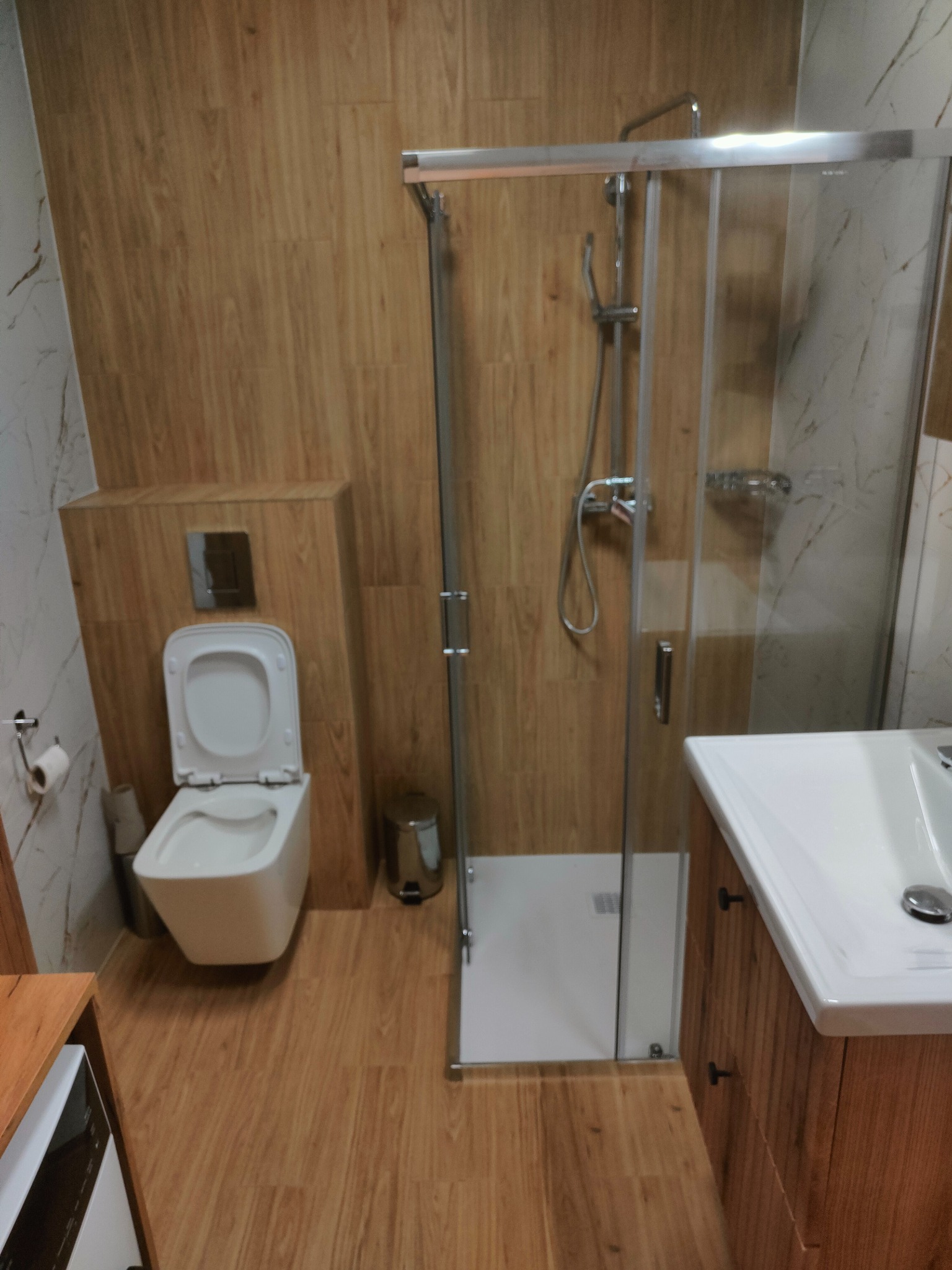 Nowoczesna łazienka z kabiną prysznicową, toaletą i umywalką w drewnianej zabudowie. Ściany wykończone płytkami imitującymi drewno i marmur.