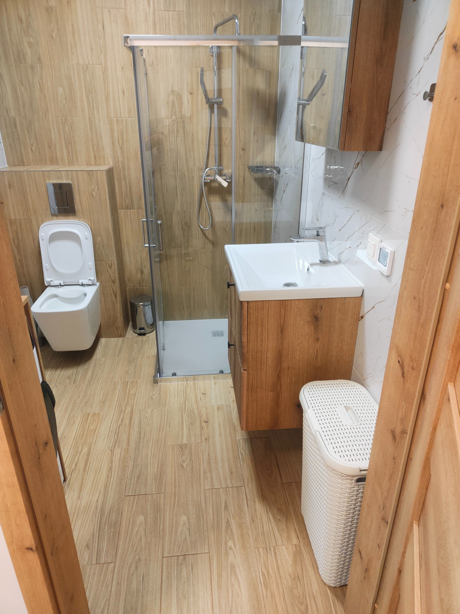 Nowoczesna łazienka w odcieniach beżu i bieli: kabina prysznicowa ze szklanymi drzwiami, umywalka z szafką w kolorze drewna i podwieszana toaleta.