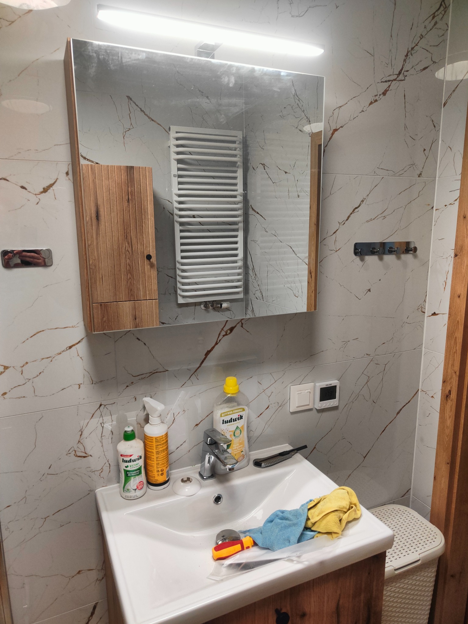 Wykończona łazienka z marmurowymi płytkami. Widoczne lustro, szafka z drewnianymi frontami, grzejnik drabinkowy i umywalka z akcesoriami.