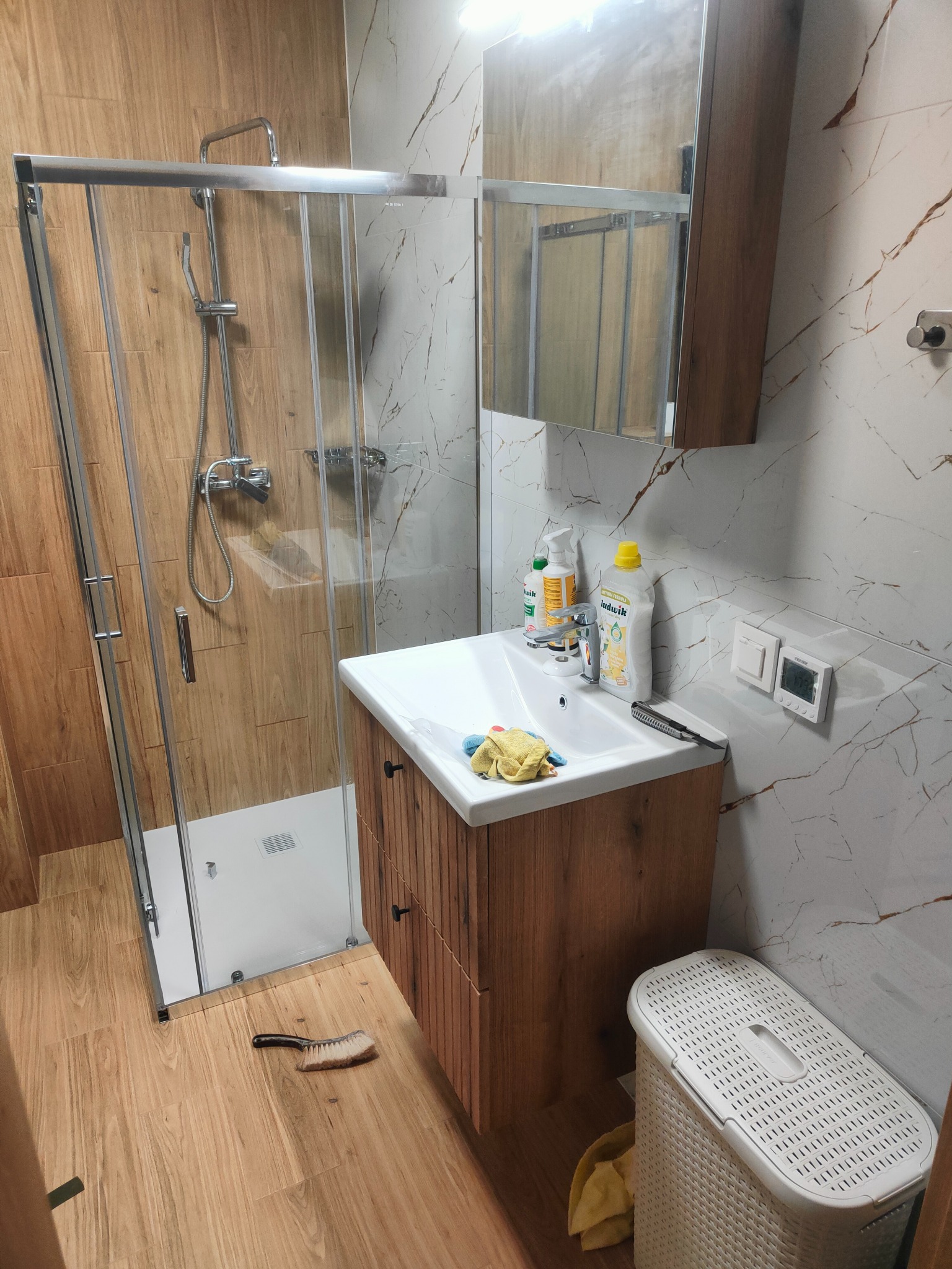 Nowoczesna łazienka z kabiną prysznicową typu walk-in, drewnianą szafką pod umywalką i marmurowymi płytkami ściennymi z brązowymi żyłami.