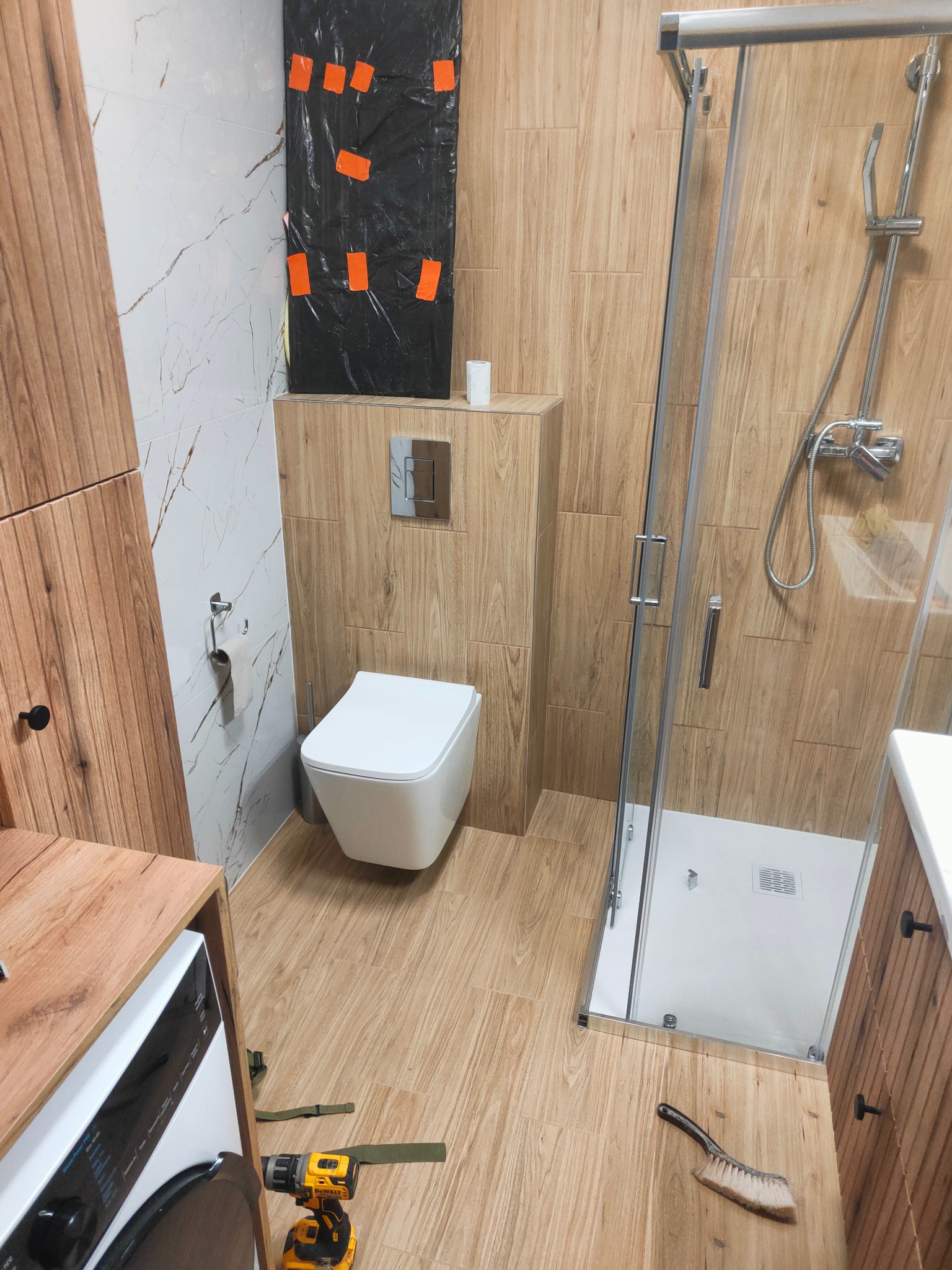 Wykończona łazienka z kabiną prysznicową i WC w zabudowie. Widoczna pralka, drewnopodobne płytki i narzędzia glazurnicze.