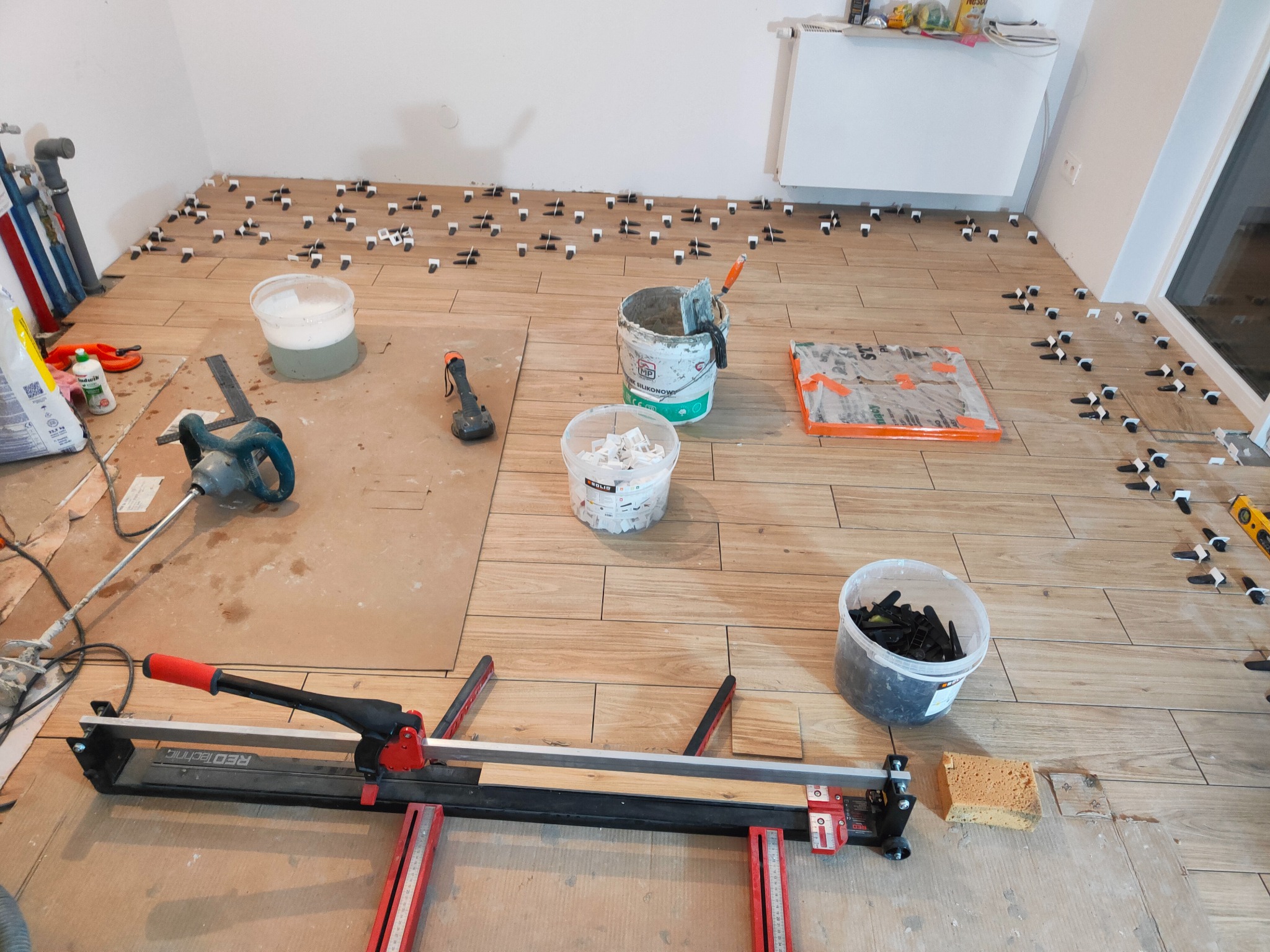Układanie płytek podłogowych z użyciem klipsów poziomujących i przecinarki RedTechnic. Widoczne narzędzia glazurnicze i materiały budowlane w jasnym pomieszczeniu.