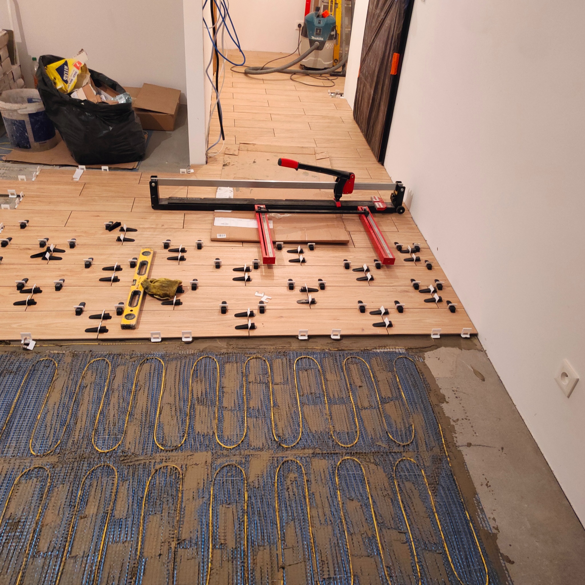 Montaż ogrzewania podłogowego i układanie płytek drewnopodobnych. Widoczna poziomica, prasa do cięcia płytek i krzyżyki dystansowe.