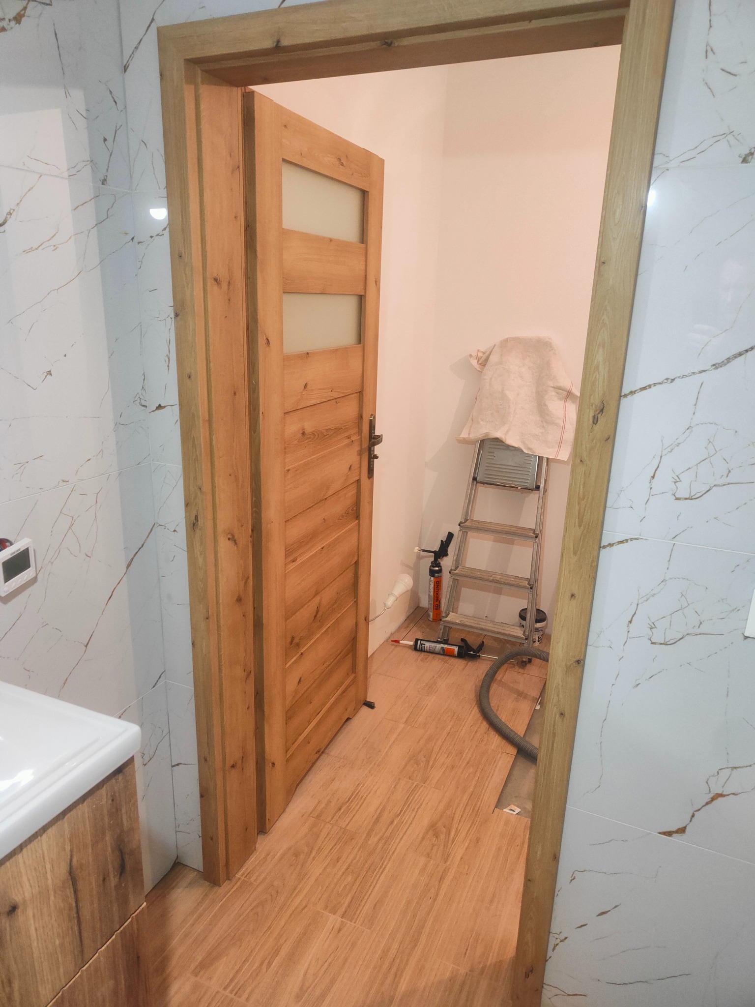 Montaż drewnianych drzwi z szybami w łazience z marmurowymi płytkami. Widoczna drabina i narzędzia.