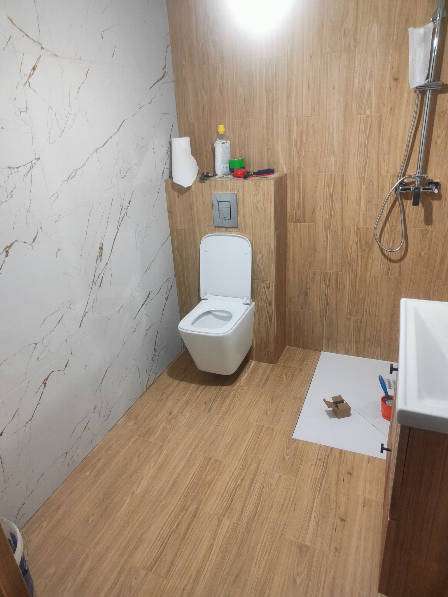 Wykończona łazienka z drewnopodobnymi i marmurowymi płytkami, wisząca toaleta, częściowo widoczna umywalka i armatura prysznicowa. W tle akcesoria glazurnicze.