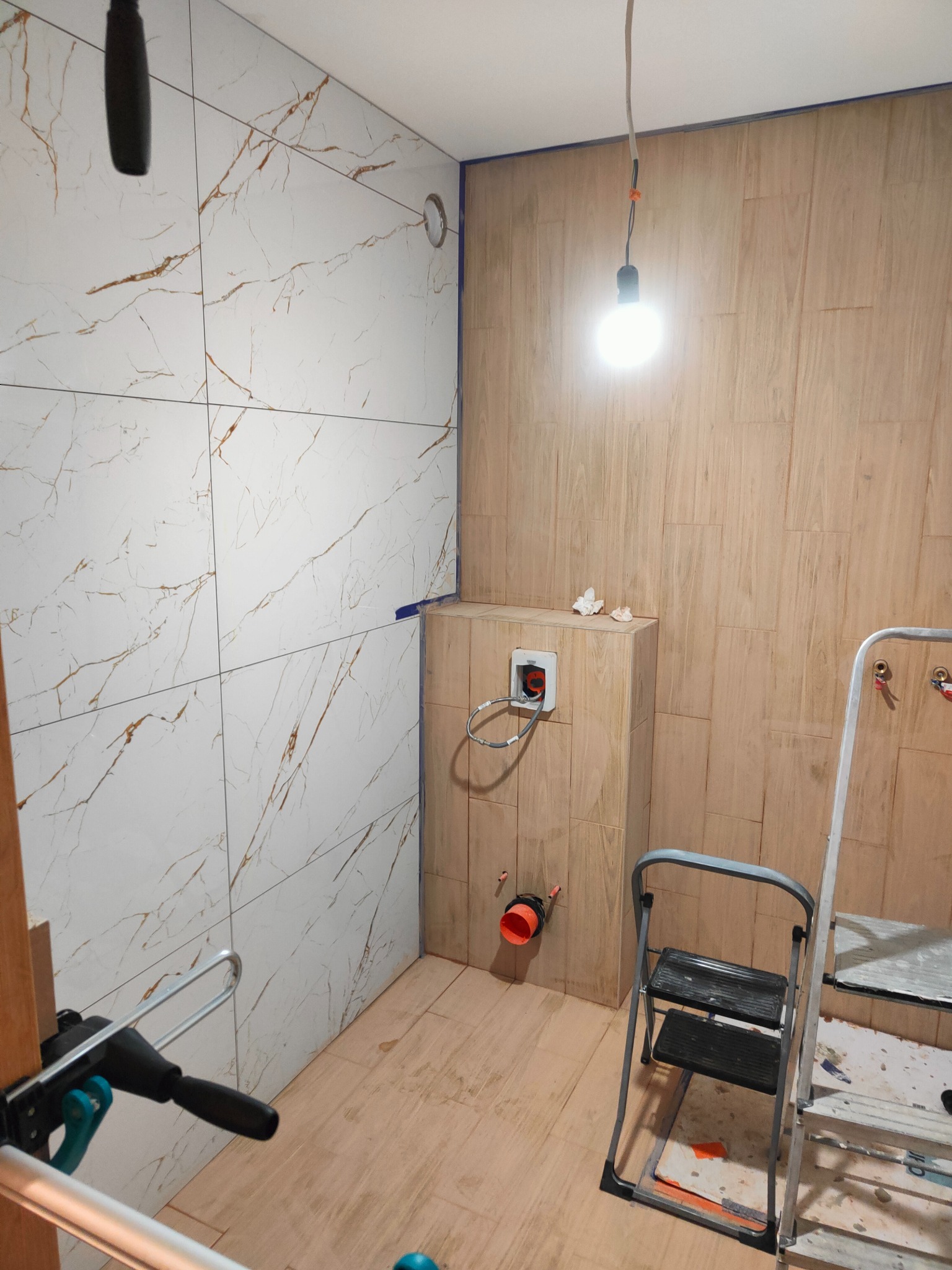 Łazienka w trakcie wykańczania: białe płytki imitujące marmur na ścianie z lewej, drewnopodobne z prawej. Widoczna drabina i narzędzia, surowa żarówka.