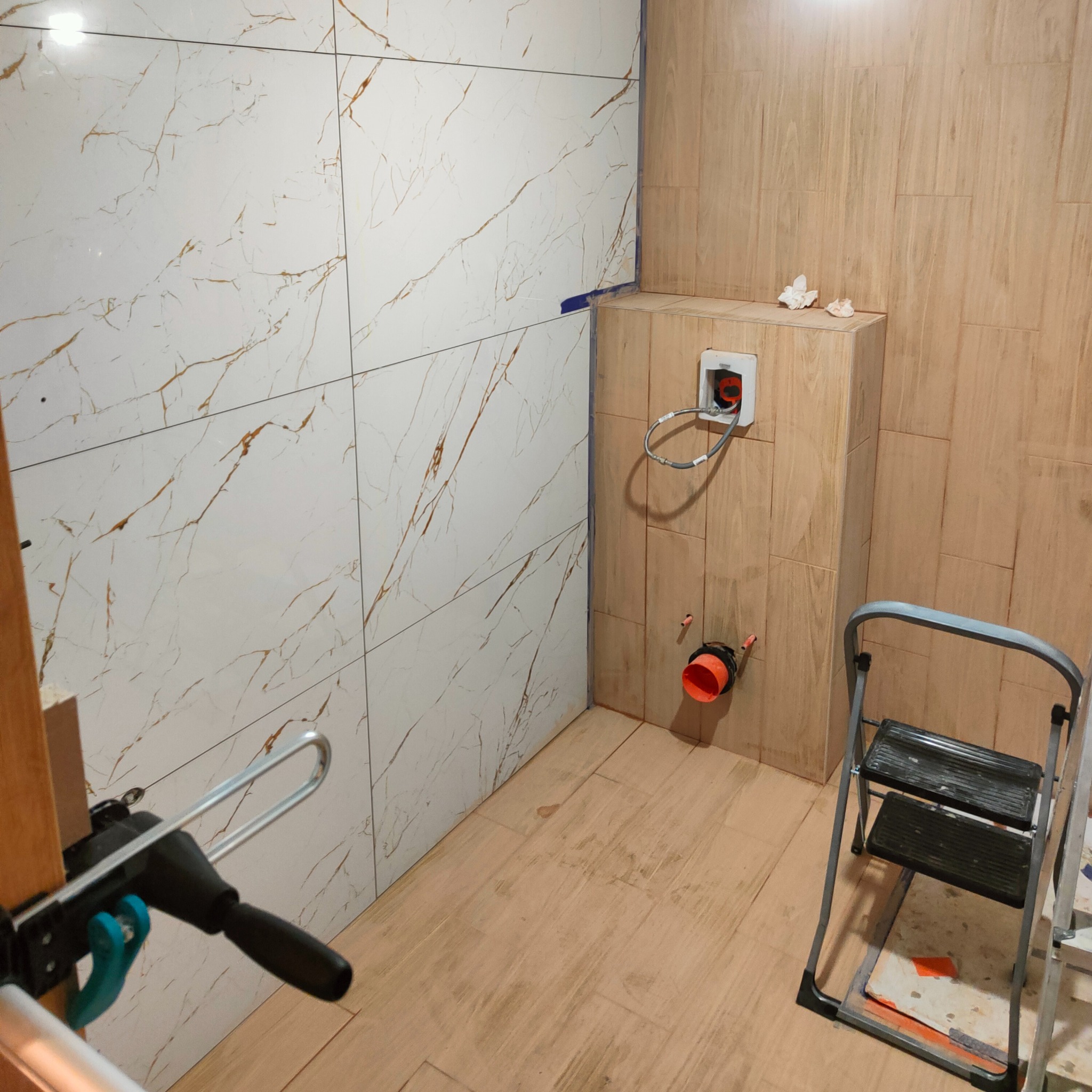 Łazienka w trakcie wykańczania: ściany częściowo wyłożone płytkami imitującymi marmur i drewno, widoczna instalacja sanitarna, drabina i narzędzia.