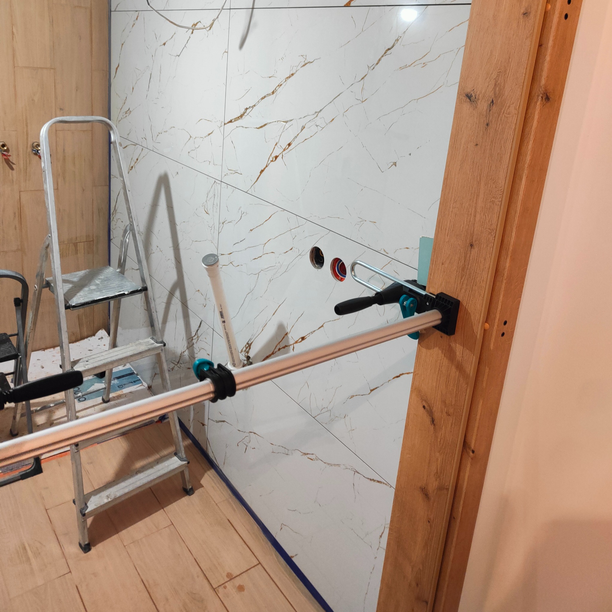 Białe płytki imitujące marmur z złotymi żyłami, zamontowane w łazience. Widoczna drabina, rury i drewniana futryna. Montaż płytek w trakcie.