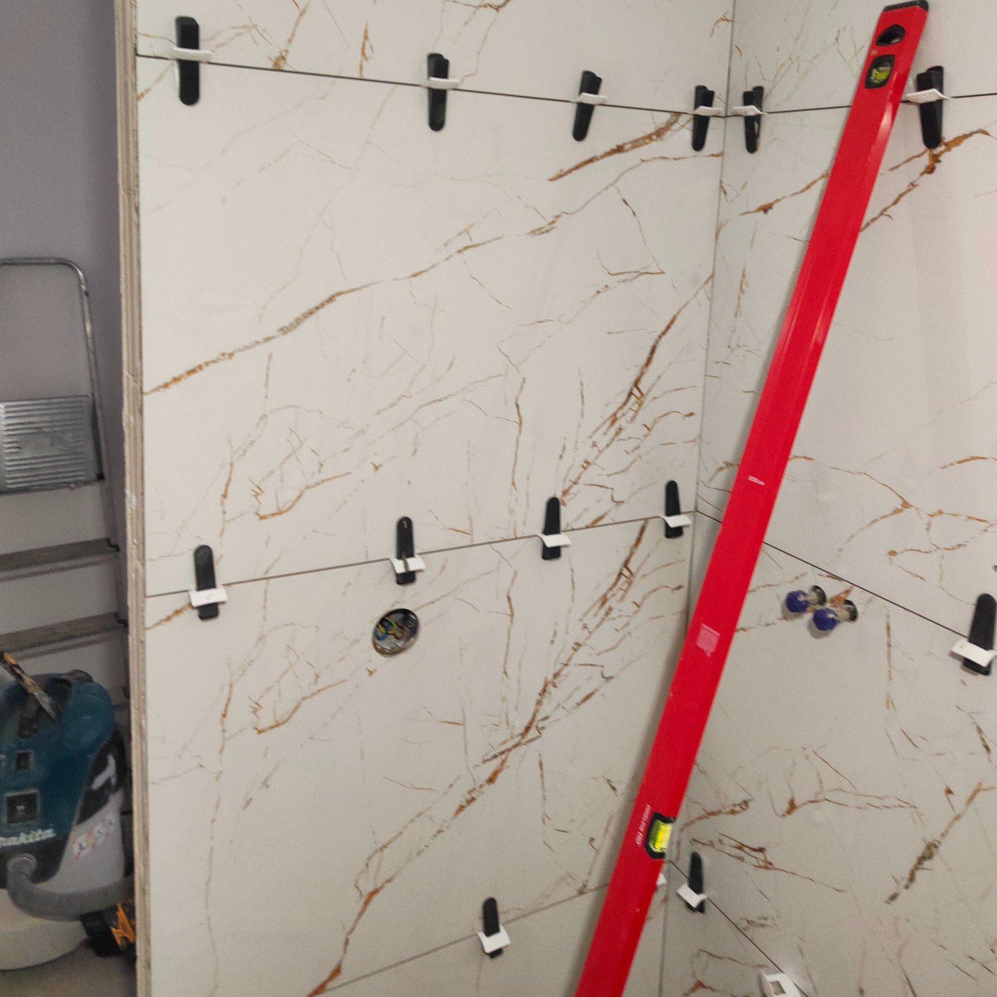 Ściana z płytek imitujących marmur, w trakcie układania. Widoczny poziomica i system poziomowania płytek. W tle drabina i odkurzacz budowlany.