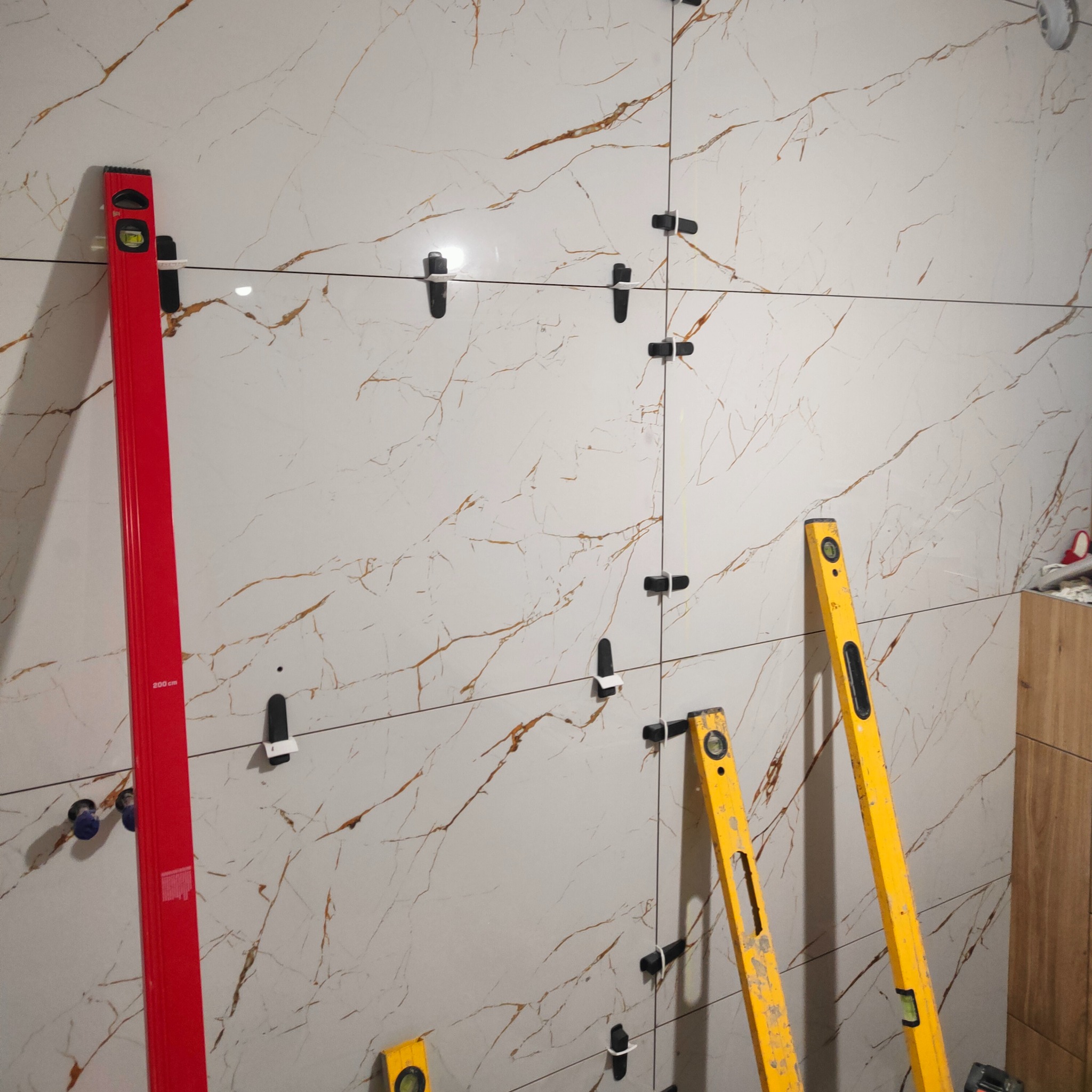 Ściana z płytek imitujących marmur, poziomowana przy użyciu poziomnic. Widoczne klipsy dystansowe. Czerwona poziomnica oparta o ścianę po lewej stronie.