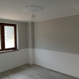 Józef Kaminski - Wnętrze pokoju po remoncie: szare ściany z białą lamperią, drewniane okno, szara podłoga z paneli, ozdobna rozeta sufitowa z wystającymi kablami.