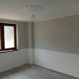 Wnętrze pokoju po remoncie: szare ściany z białą lamperią, drewniane okno, szara podłoga z paneli, ozdobna rozeta sufitowa z wystającymi kablami.