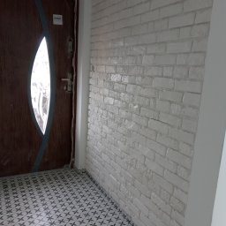 Józef Kaminski - Wnętrze po remoncie: brązowe drzwi z owalnym przeszkleniem, ściana wyłożona białą cegłą dekoracyjną oraz podłoga z płytek w geometryczny wzór.