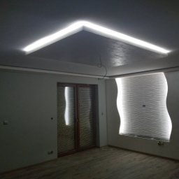 Józef Kaminski - Nowoczesne wnętrze pokoju z podwieszanym sufitem z oświetleniem LED, dekoracyjną ścianą z efektem fal i drewnianą stolarką okienną.