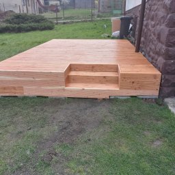 Józef Kaminski - Nowo wybudowany drewniany taras z małymi schodkami, przylegający do kamiennej ściany budynku, otoczony trawnikiem.