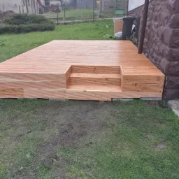 Nowo wybudowany drewniany taras z małymi schodkami, przylegający do kamiennej ściany budynku, otoczony trawnikiem.