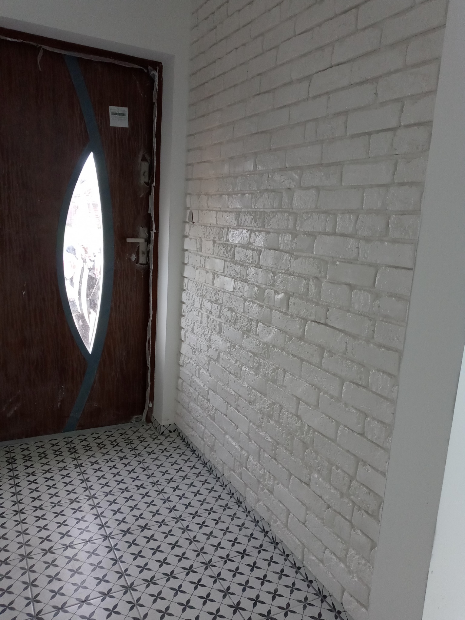 Wnętrze po remoncie: brązowe drzwi z owalnym przeszkleniem, ściana wyłożona białą cegłą dekoracyjną oraz podłoga z płytek w geometryczny wzór.