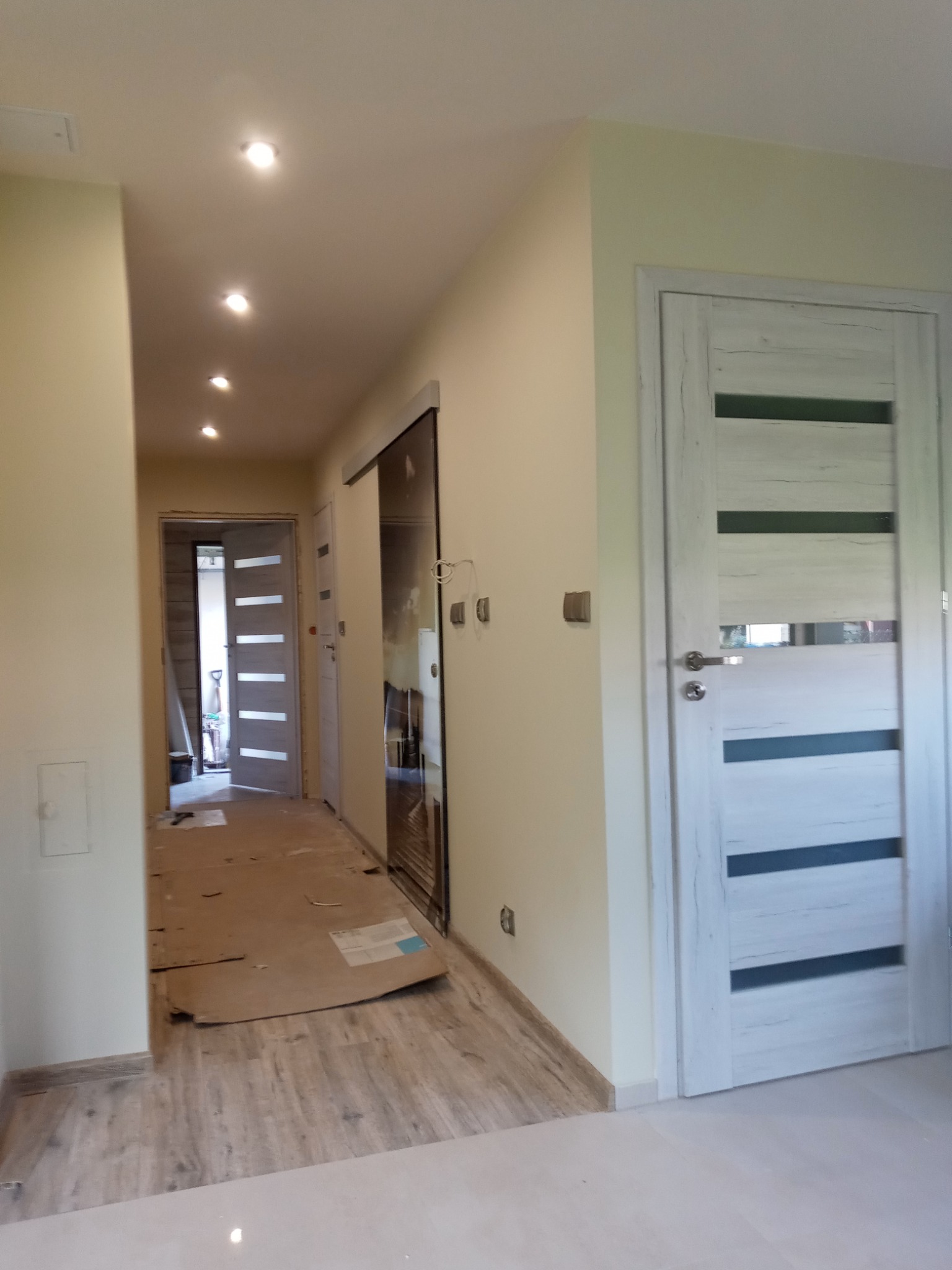 Korytarz w trakcie remontu z zamontowanymi drzwiami wewnętrznymi z poziomymi przeszkleniami, podłoga zabezpieczona kartonem, widoczne oświetlenie sufitowe.