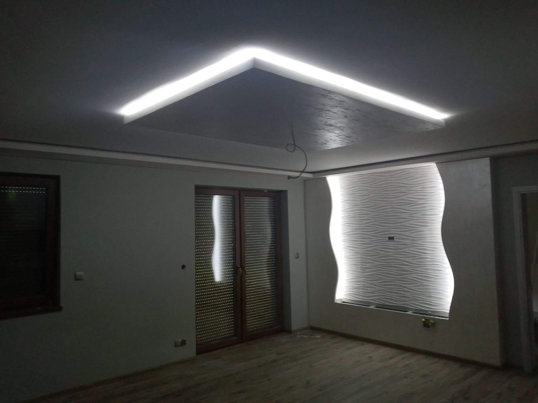 Nowoczesne wnętrze pokoju z podwieszanym sufitem z oświetleniem LED, dekoracyjną ścianą z efektem fal i drewnianą stolarką okienną.