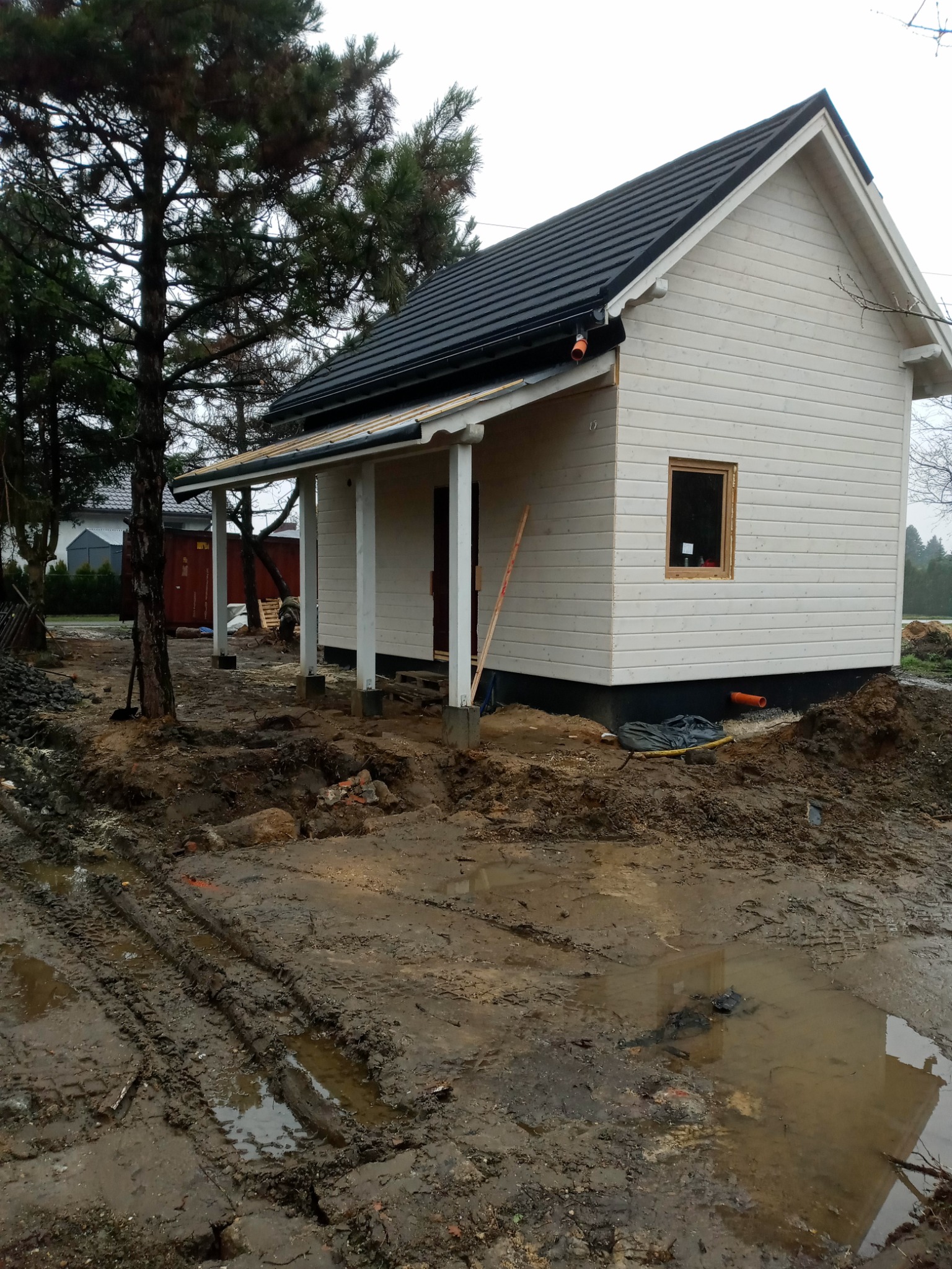 Surowy stan budowy małego, parterowego domu z białego drewna z czarnym dachem, widoczne ślady opon i błoto na placu budowy, drzewa w tle.