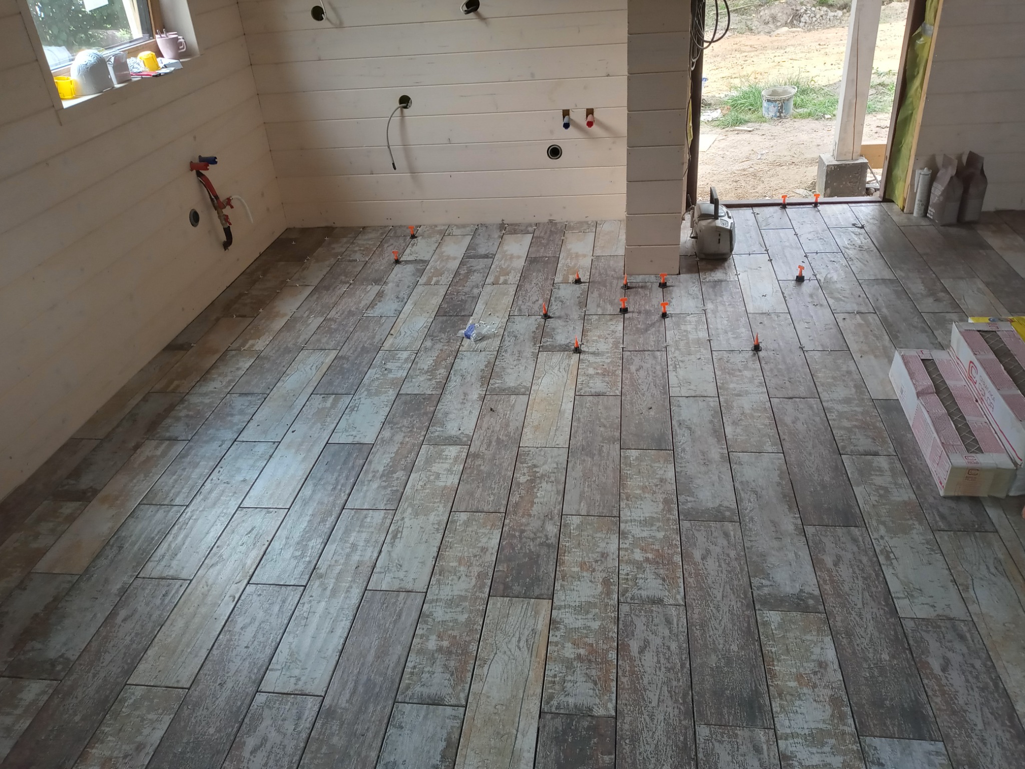 Wnętrze domu w trakcie wykańczania: ułożona podłoga z płytek imitujących drewno, z pomarańczowymi elementami poziomującymi, widoczne przygotowane ściany z drewna i instalacje.