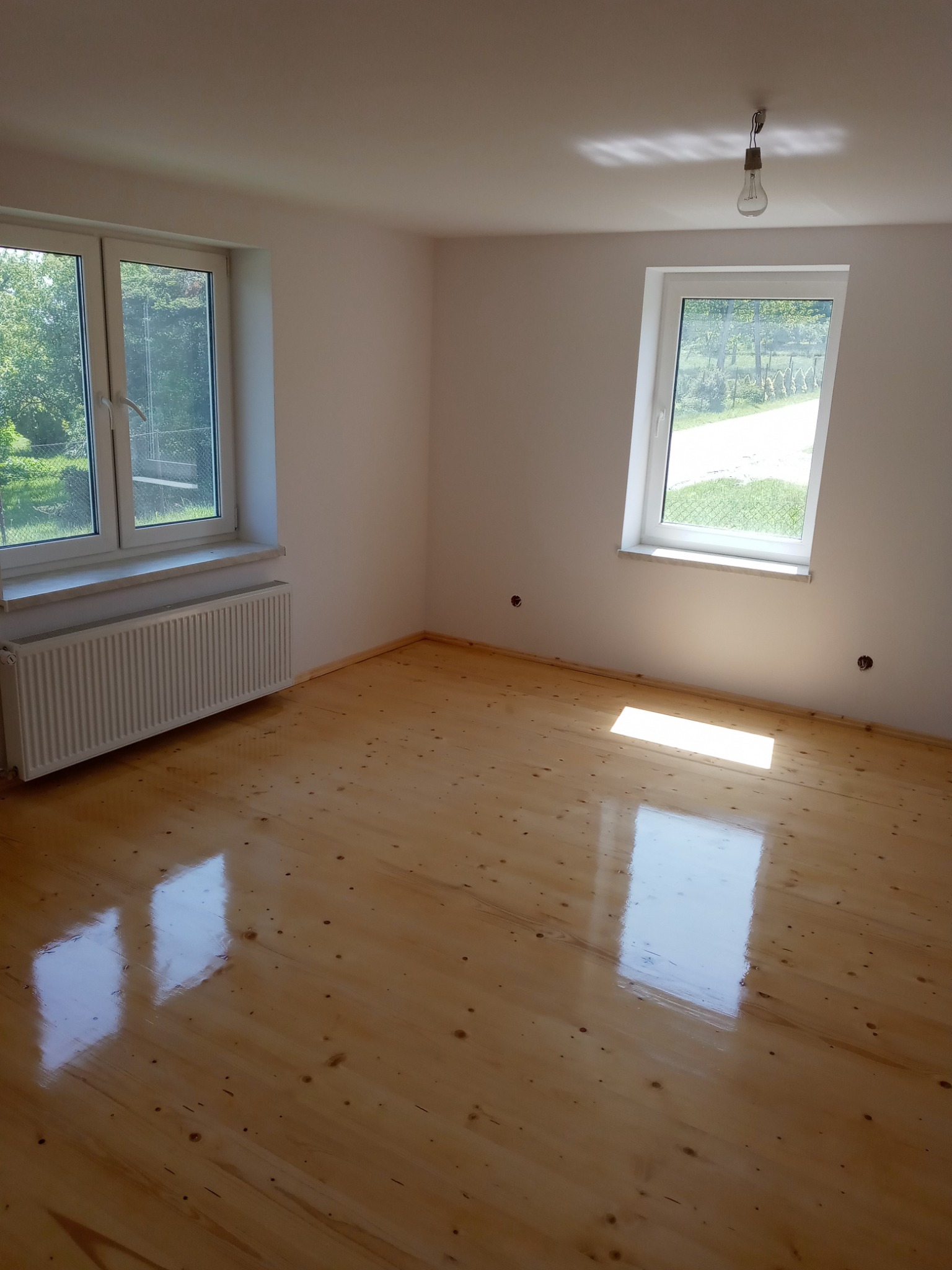 Pusty, jasny pokój po renowacji z lakierowaną, drewnianą podłogą, białymi ścianami, dwoma oknami z widokiem na zieleń i grzejnikiem pod jednym z nich. Na suficie wisi żarówka na kablu.