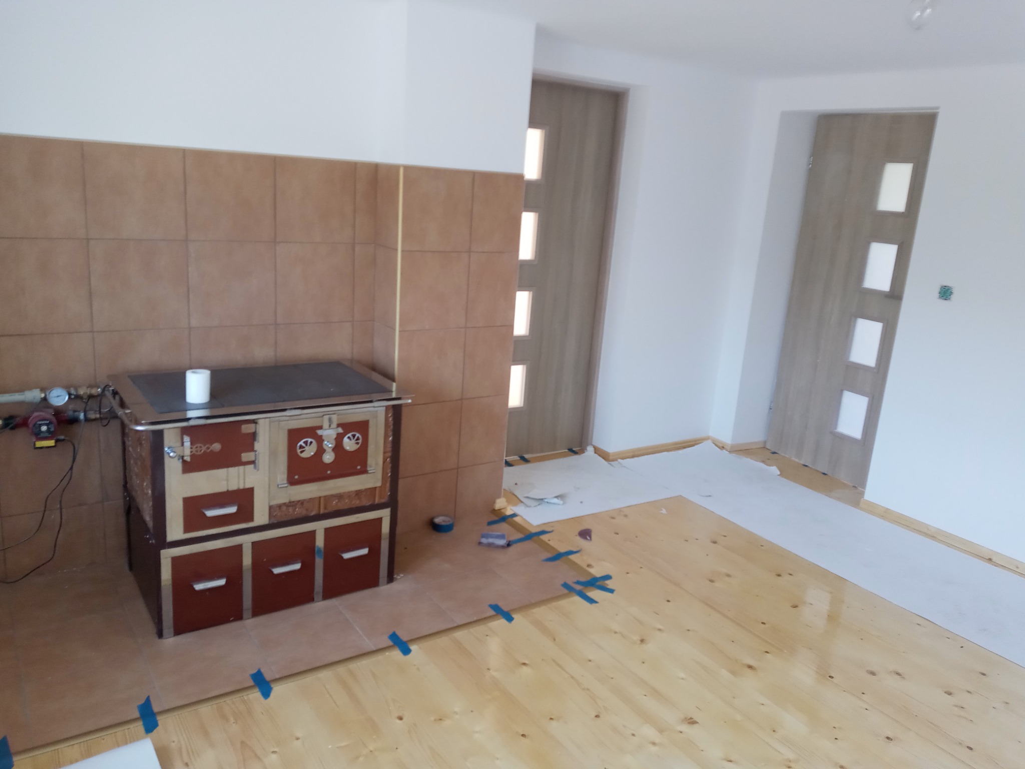 Fragment wnętrza w trakcie remontu: widoczny piec kaflowy z podłączeniem do instalacji, częściowo wykończona ściana z brązowych płytek, surowa drewniana podłoga zabezpieczona taśmą malarską...