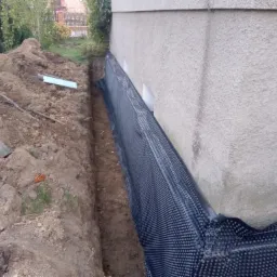 Przygotowanie do izolacji fundamentów: wykop, folia kubełkowa przy ścianie budynku, widoczny fragment styropianu.