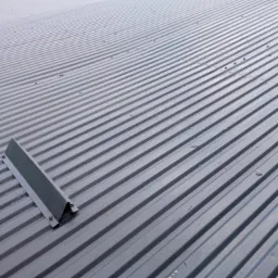 Szare, pomalowane blachy trapezowe tworzące pokrycie dachowe z widocznym elementem mocującym w lewym górnym rogu, sfotografowane pod kątem.