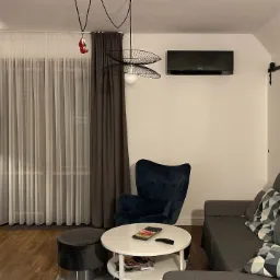 Czarny klimatyzator Mitsubishi Electric zamontowany na ścianie w nowoczesnym salonie z szarą sofą, welurowym fotelem i designerską lampą wiszącą.