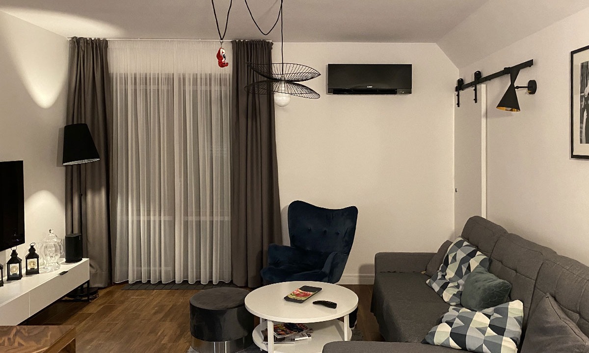 Czarny klimatyzator Mitsubishi Electric zamontowany na ścianie w nowoczesnym salonie z szarą sofą, welurowym fotelem i designerską lampą wiszącą.