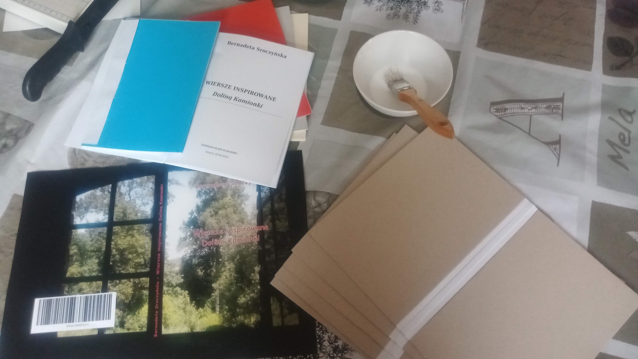 Introligatornia: książki w trakcie oprawy, pędzel w miseczce z klejem, okładki i narzędzia na wzorzystym obrusie. Widoczne etapy pracy introligatorskiej.