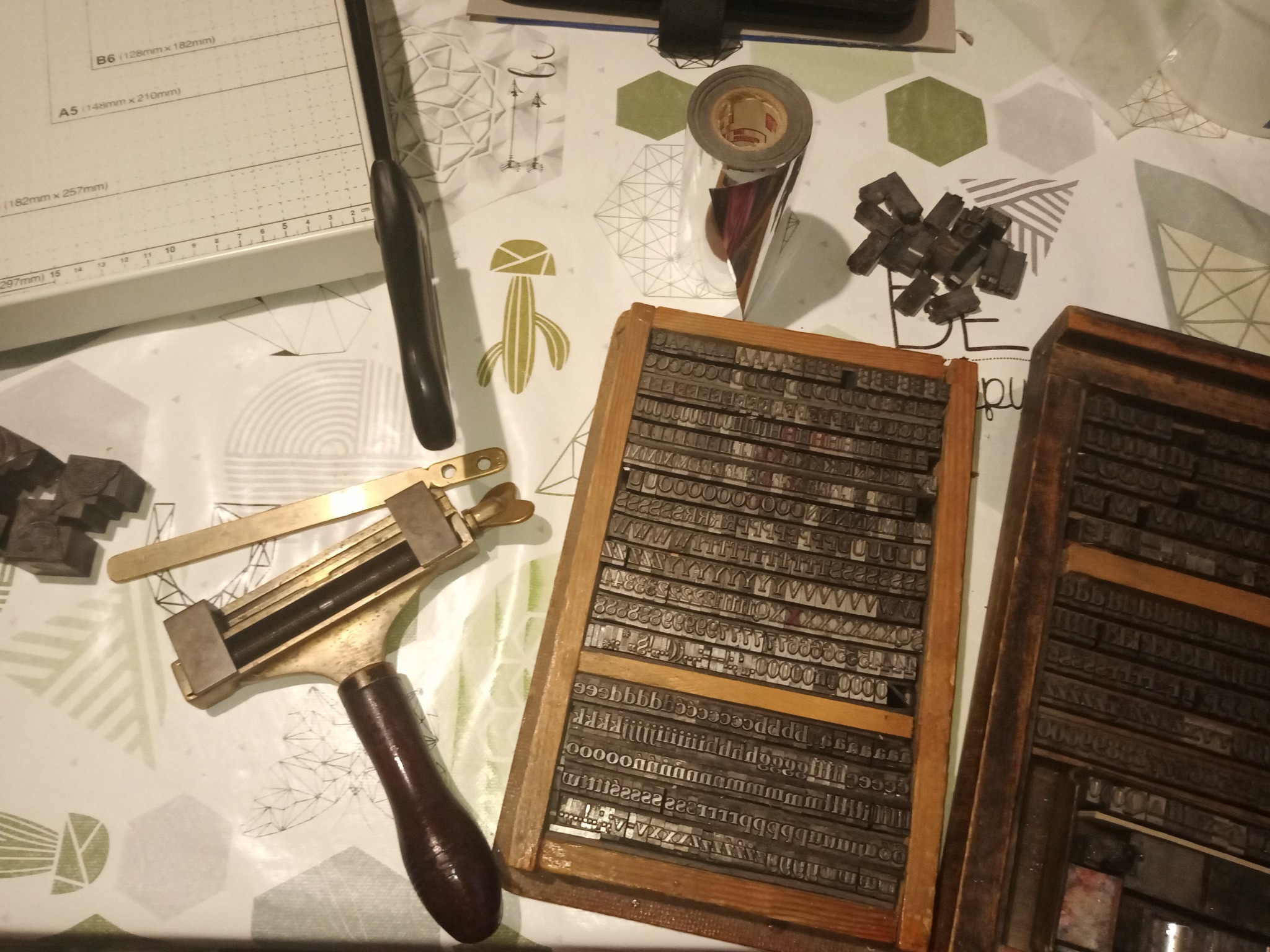 Przygotowane do użycia czcionki drukarskie w drewnianych kasetach, obok złocarki z brązową rączką, noża do papieru i rolki folii do hot stampingu na stole z wzorzystym obrusem.