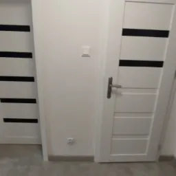 Białe drzwi wewnętrzne z czarnymi poziomymi paskami, widoczne gniazdko elektryczne i listwa przypodłogowa, szara podłoga. Minimalistyczny design.