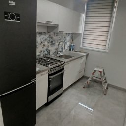 REM-TUR - Nowoczesna kuchnia z czarną lodówką Beko, białymi szafkami i kwiatowym backsplash'em. Metalowa drabina stoi obok okna z żaluzjami, oświetlona lampą sufitową.