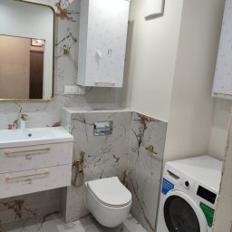 REM-TUR - Elegancka łazienka z marmurowymi płytkami, złotymi akcentami i nowoczesną armaturą. Wisząca toaleta, szafka nad nią i pralka tworzą spójną całość.