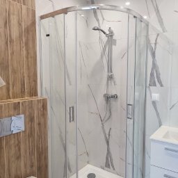 REM-TUR - Nowoczesna kabina prysznicowa z marmurowymi płytkami i drewnianymi akcentami. Widoczna armatura, biała umywalka i toaleta. Minimalistyczny design, oświetlenie sufitowe.