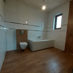 REM-TUR - Wykończona łazienka z drewnopodobną podłogą, białą armaturą i płytkami. Widoczna wanna z dekoracyjnym frontem i toaleta podtynkowa. Minimalistyczny styl.