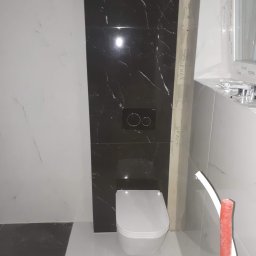 REM-TUR - Nowoczesna łazienka w trakcie wykończenia: biała ceramika sanitarna na tle czarnych płytek imitujących marmur. Widoczne rury instalacyjne i fragment okna.