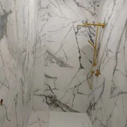 Wykończona łazienka z marmurowymi płytkami i złotą armaturą prysznicową. Widoczne niedokończone prace elektryczne.