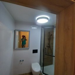 REM-TUR - Nowoczesna, jasna łazienka z kabiną prysznicową i podświetlanym lustrem. Białe płytki na ścianach, minimalistyczna toaleta, ciemna rama kabiny.