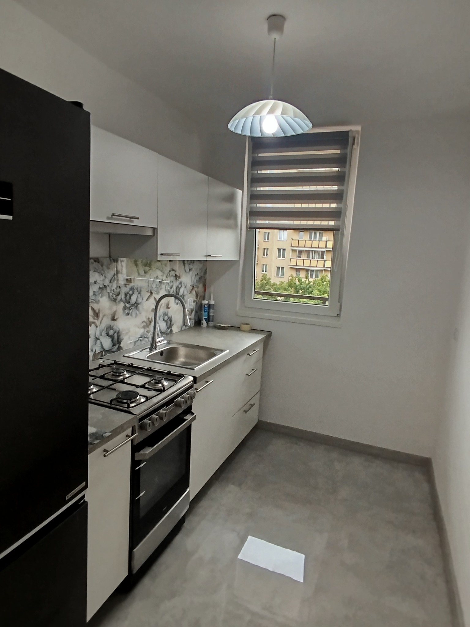 Jasna kuchnia z kwiatowym motywem na ścianie, widok na blok zza okna, nowoczesne oświetlenie sufitowe i szare płytki podłogowe. Ujęcie z perspektywy.