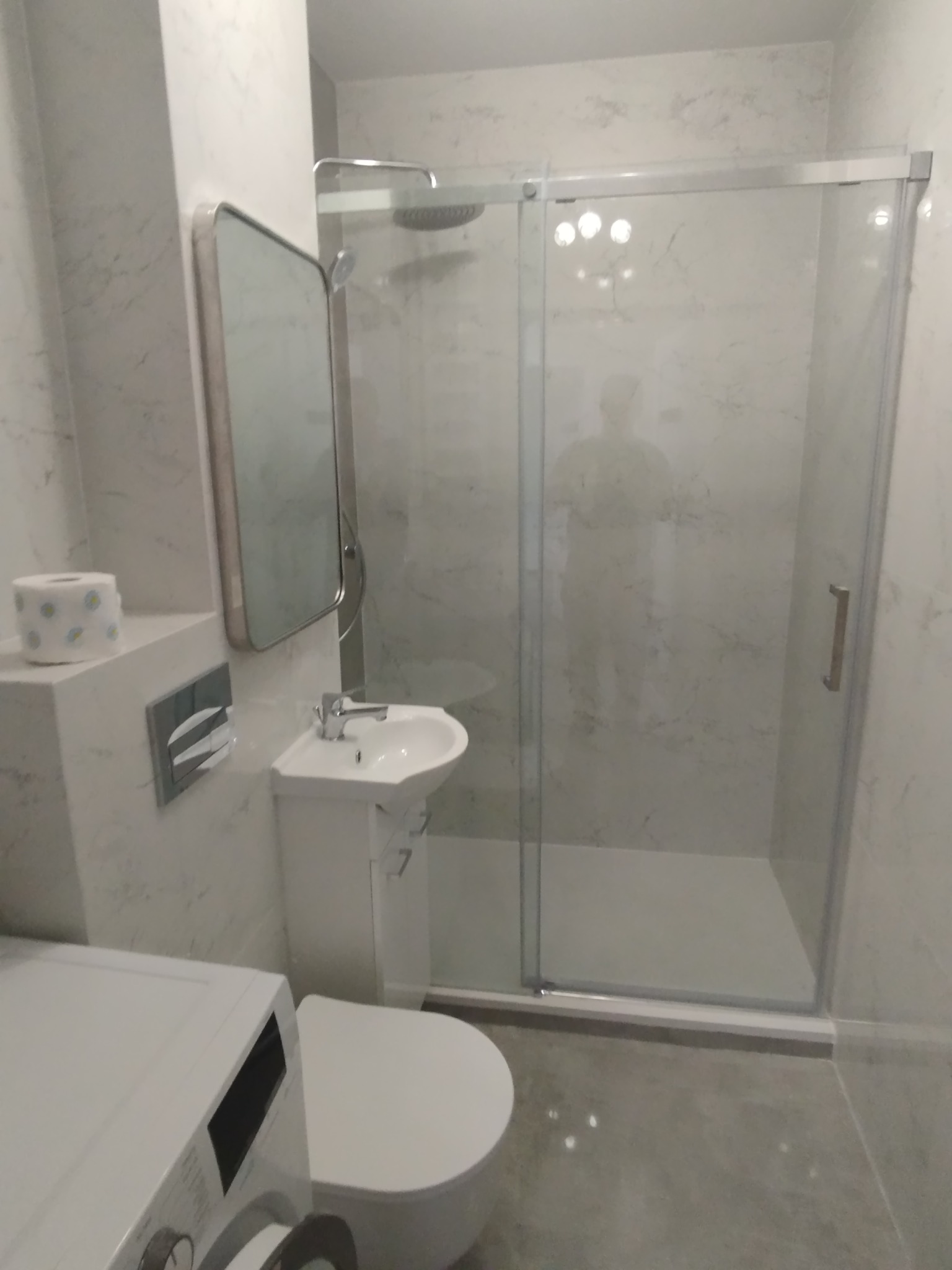 Nowoczesna łazienka z kabiną prysznicową typu walk-in, umywalką nablatową i toaletą. Ściany wykończone marmurkową okładziną. Widoczna pralka w dolnym rogu.