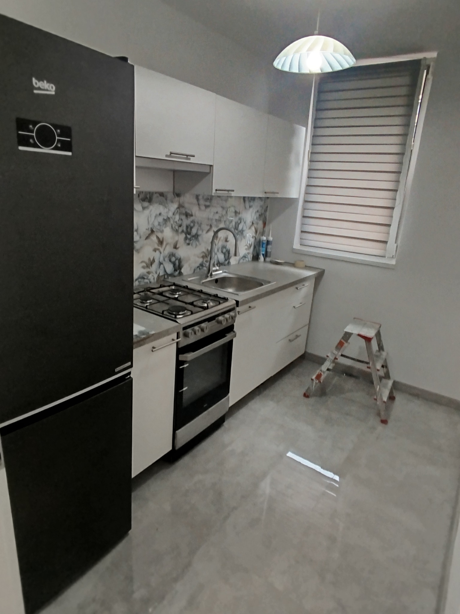 Nowoczesna kuchnia z czarną lodówką Beko, białymi szafkami i kwiatowym backsplash'em. Metalowa drabina stoi obok okna z żaluzjami, oświetlona lampą sufitową.