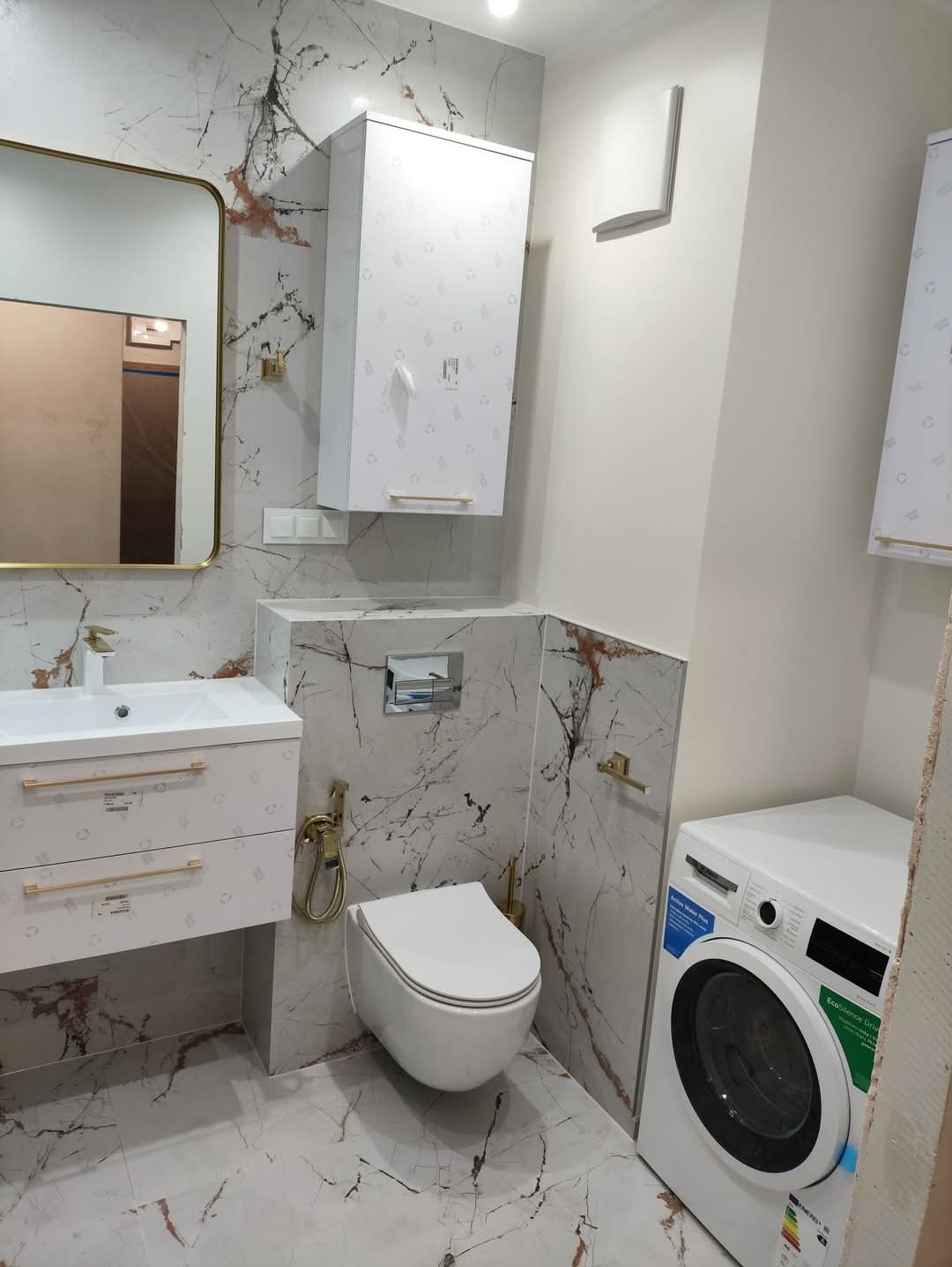 Elegancka łazienka z marmurowymi płytkami, złotymi akcentami i nowoczesną armaturą. Wisząca toaleta, szafka nad nią i pralka tworzą spójną całość.