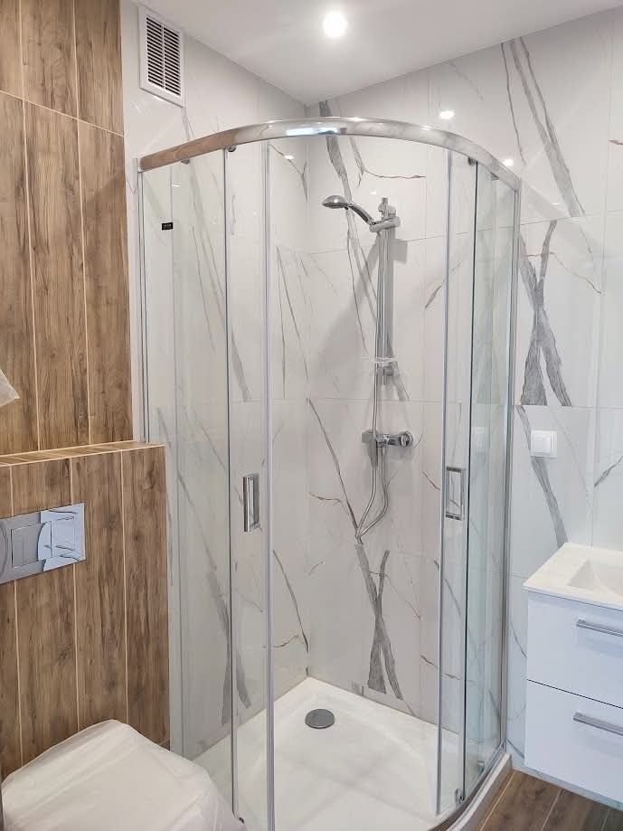 Nowoczesna kabina prysznicowa z marmurowymi płytkami i drewnianymi akcentami. Widoczna armatura, biała umywalka i toaleta. Minimalistyczny design, oświetlenie sufitowe.