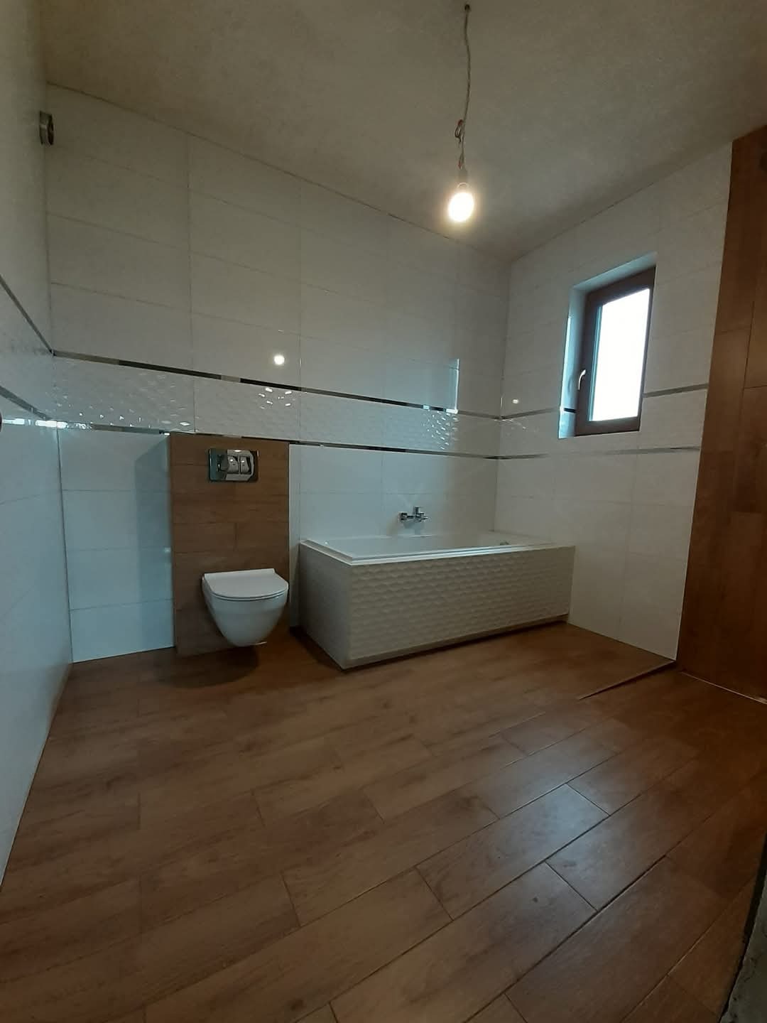 Wykończona łazienka z drewnopodobną podłogą, białą armaturą i płytkami. Widoczna wanna z dekoracyjnym frontem i toaleta podtynkowa. Minimalistyczny styl.