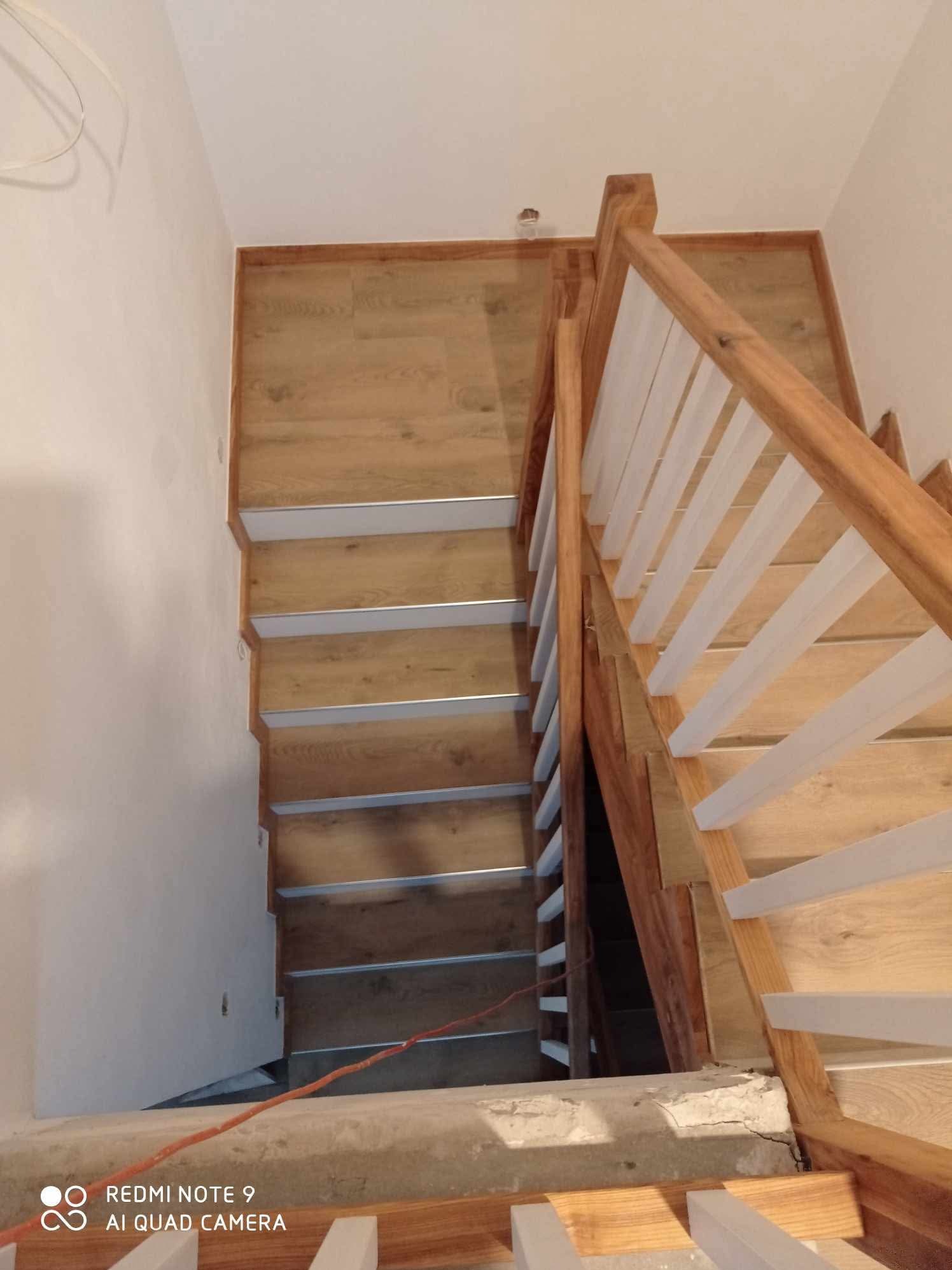 Drewniane schody z białymi balustradami i metalowymi listwami na stopniach, widok z góry. Nowoczesne wykończenie, jasne kolory, ukończony remont.