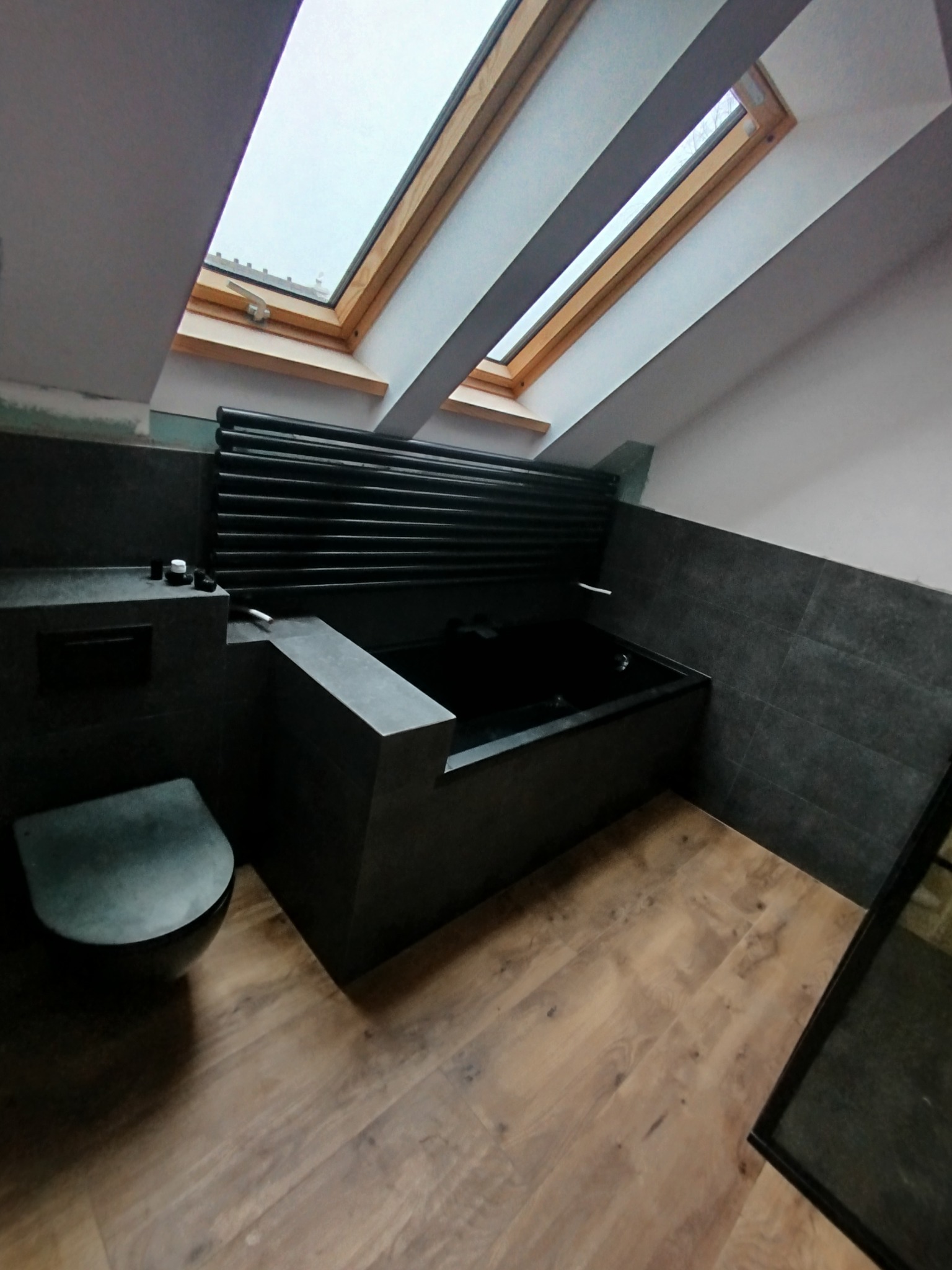 Nowoczesna, ciemna łazienka na poddaszu z oknem dachowym, drewnianą podłogą i czarną armaturą. Uwagę przykuwa wanna obudowana płytkami w kolorze grafitu.