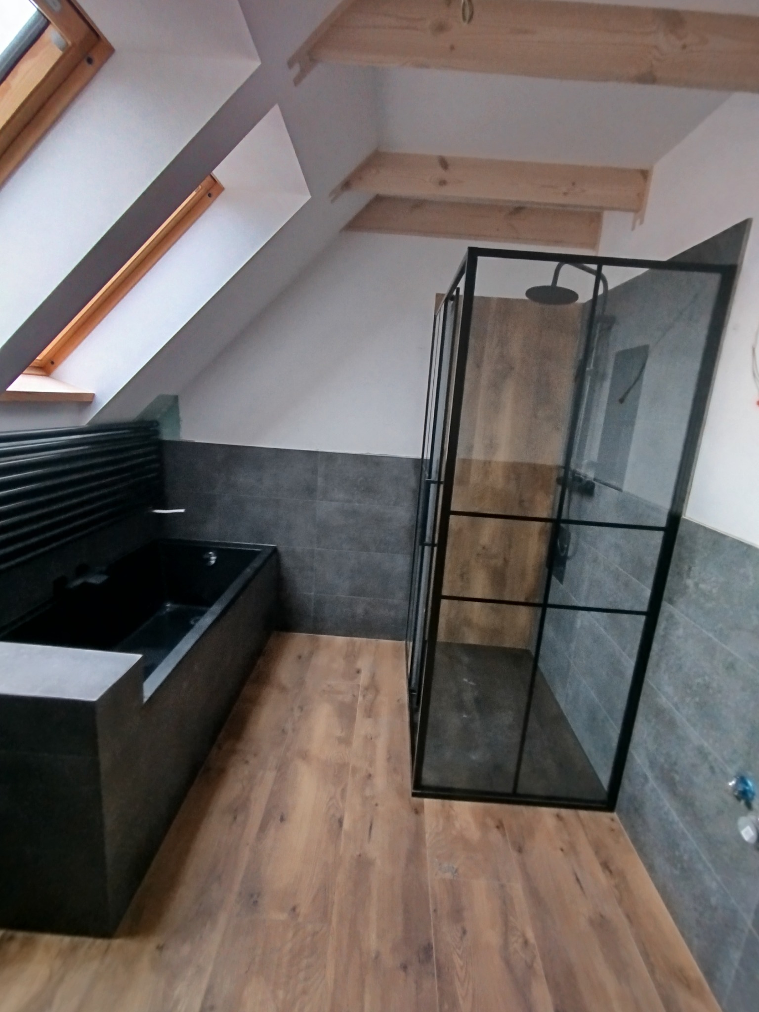 Nowoczesna łazienka na poddaszu: czarna wanna z grzejnikiem drabinkowym i kabina prysznicowa typu walk-in z czarnymi szprosami, drewnopodobna podłoga, szare płytki.